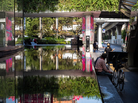 This photo showcases a reflective pool outside the Emporium Mall, located by Phrom Phong station along Sukhumvit Road in Bangkok, Thailand. In the photo, people wearing face masks can be seen enjoying the outdoor space while maintaining social distance. Tのeditorial素材