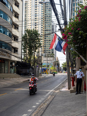 This photograph captures the essence of Bangkok - the bustling yet serene streets, the ubiquitous motorcycle taxis, and the proud waving of the Thai flag. The image of a hotel guard standing watch amidst the emptiness adds an intriguing dimension to the sのeditorial素材