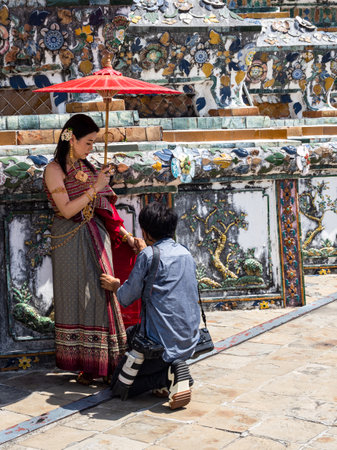 A beautiful Thai woman adorned in traditional clothing holds a red umbrella as a photographer prepares to capture her elegance in front of the stunning Wat Arun Temple, showcasing the rich cultural heritage and beauty of Thailand.のeditorial素材