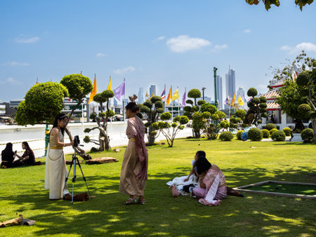 Thai local women wearing traditional clothing gather on a grassy lawn with a photo project by the Wat Arun Temple next to the Chao Praya River with the Bangkok skyline visible in the distance. It's a beautiful representation of the intersection of cultureのeditorial素材