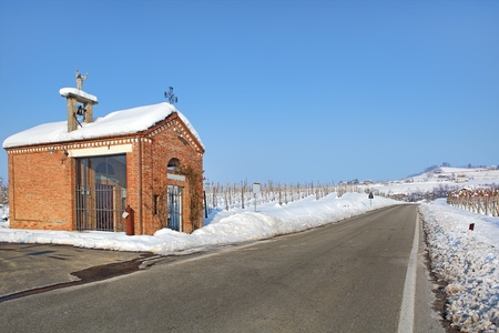 Small red chapel on the roadside among vineyards covered by white snow under clear blue winter sky in Piedmont, Northern Italy.の写真素材