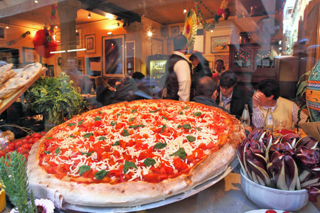 VENICE - MARCH 04: Big round plate of pizza with red tomatoes and cheese displayed on restaurant window and visitors inside on background. Pizza is traditional italian dish very popular around the world in Venice, Italy on March 04, 2012.のeditorial素材