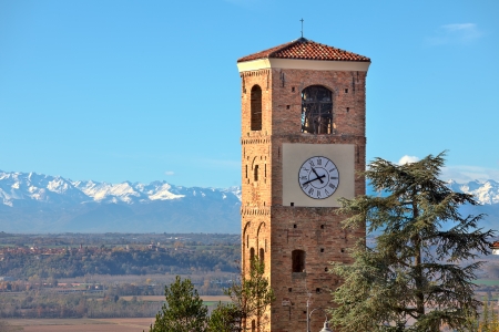 View on old brick belfry tower with big clock and mountains with snowy peaks on background under blue sky in Piedmont, Northern Italy の写真素材