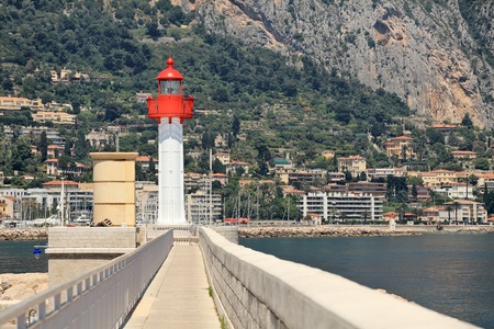 White and red lighthouse at the end of narrow promenade along the sea as houses and hotels on the slope on background in Menton, France の写真素材