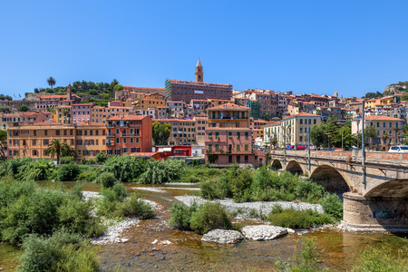 Urban bridge across riverbed overgrown with trees, shrubs and grass and old town of Ventimiglia under blue sky in Italy の写真素材