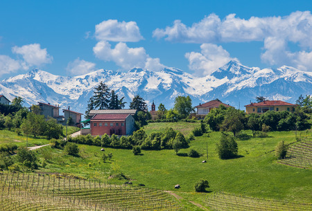 Rural houses on green field and snowy mountains on background in spring in Piedmont, Italy の写真素材
