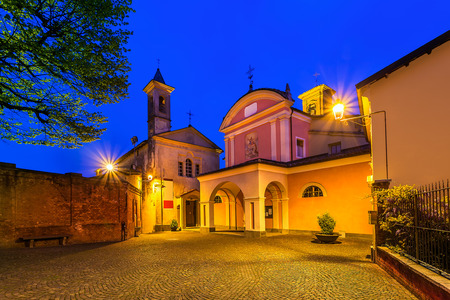 Small cobbled town square and two churches illuminated by lamppost early in the morning in Piedmont, Italy.の写真素材