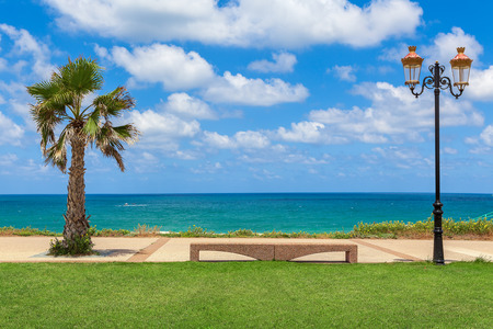 Stone bench between palm and decorative lamppost with beautiful view on Mediterranean sea under blue sky with white clouds in Israel.の写真素材