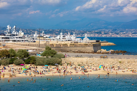 ANTIBES, FRANCE - SEPTEMBER 04, 2018: People on Plage de la Gravette - famous and popular sand beach near the Old Town as marina and luxury yachts on background in Antibes, France.のeditorial素材
