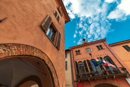 Old historic house and town hall with balcony, flags and clock under beautiful sky in Alba, Piedmont, Northern Italy (low angle view).の写真素材