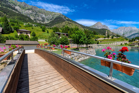 Narrow wooden bridge over small artificial lake as mountains under blue sky on background in town of Pietraporzio in Piedmont, Northern Italy.の写真素材