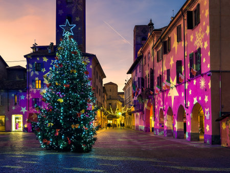 A tall Christmas tree with sparkling lights stands in the center of historic Italian town square at twilight as colorful projections illuminate the surrounding buildings.の写真素材