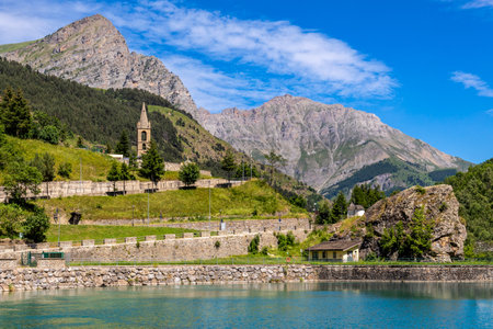 View of alpine lake, parish church on the hill and mountains under blue sky on background in small town of Pietraporzio, Italy.の写真素材