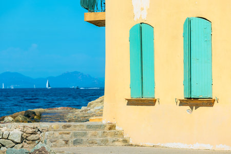 A yellow walled building with vibrant turquoise shutters overlooks the azure waters of Saint-Tropez, France.の写真素材