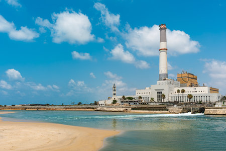 The Reading power station with tall chimney stands in the background of a sandy beach and the Mediterranean sea under a blue sky with white clouds in Tel Aviv, Israel.の写真素材