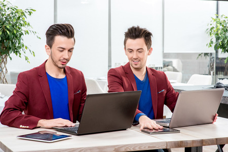 Two brothers twins in red jackets working with laptops and smart phones at the office. Family businessの写真素材