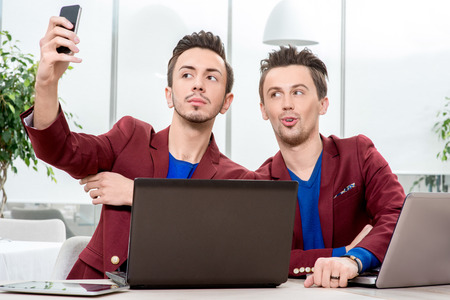 Friendly brothers twins having fun taking selfie photo with smart phone in the white home or restaurant interiorの写真素材