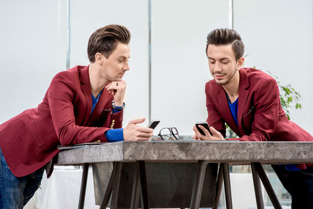 Two brothers twins in red jackets discussing something with smart phones at the office tableの写真素材