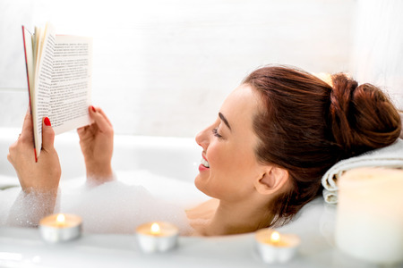 Young woman reading book while lying in the bath with foam and candlesの写真素材