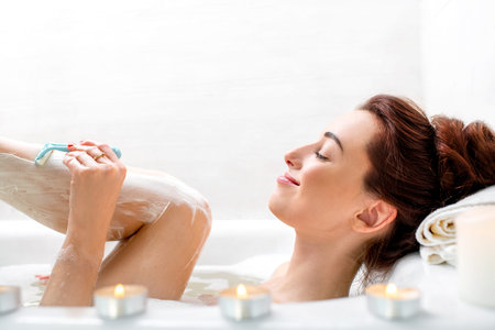 Young and positive woman shaving her legs in the bath with foam and candles.の写真素材