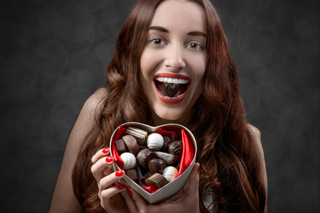 Happy and joyful young woman holding heart box with candies and eating them on grey background. Happy valentines gift conceptの写真素材