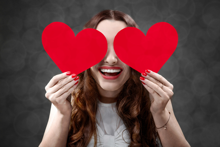 Happy and joyful young woman holding red hearts on grey background in studio. Happy valentines greeting conceptの写真素材