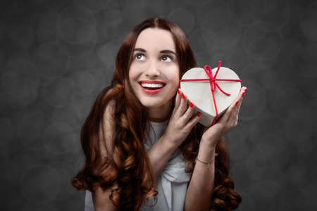 Happy and smiling young woman holding heart box with candies on grey background. Happy valentines gift conceptの写真素材