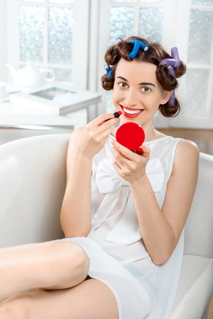 Smiling woman with hair curlers using lipstick on white domestic backgroundの写真素材