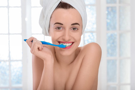 Smiling young woman with white towel on the head with healthy teeth and beautiful smile brushing her teeth in the bathroomの写真素材