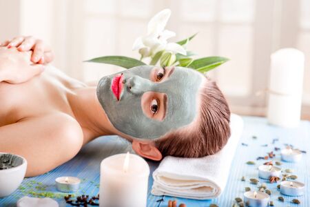 Young woman with spa facial mask on her face lying on blue table with flower, candles and sea salt in the beaty salon. Side viewの写真素材