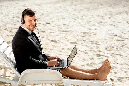 Businessman dressed in suit and shorts having video call with laptop on the sunbed at the beachの写真素材