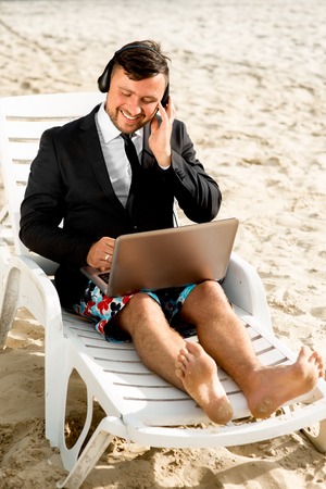 Businessman dressed in suit and shorts having video call with laptop on the sunbed at the beachの写真素材