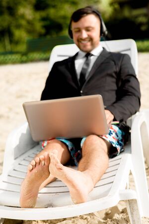 Businessman dressed in suit and shorts working with laptop on the sunbed at the beachの写真素材