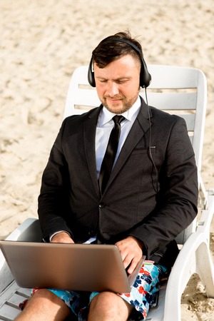 Businessman dressed in suit and shorts having video call with laptop on the sunbed at the beachの写真素材