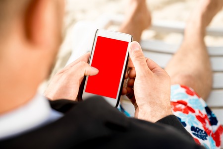 Businessman holding cellphone with empty screen sitting on the sunbed dressed in suit and shorts on the beachの写真素材