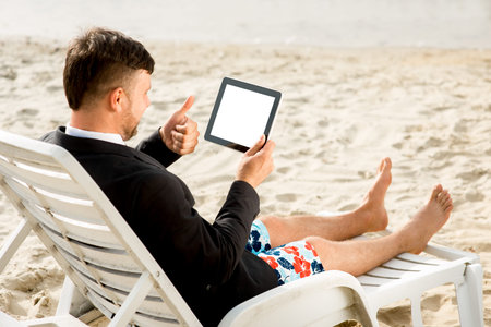 Businessman dressed in suit and shorts having video call with digital tablet on the sunbed at the beachの写真素材