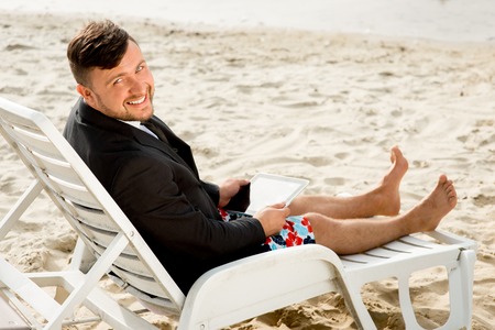 Businessman dressed in suit and shorts working with laptop on the sunbed at the beachの写真素材