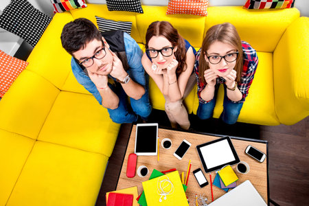 Three nerds in eyeglasses sitting on the couch and looking at camera with different gadgets on the backgroundの写真素材