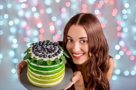Beautiful girl holding happy birthday cake and smiling looking at camera on festive light backgroundの写真素材