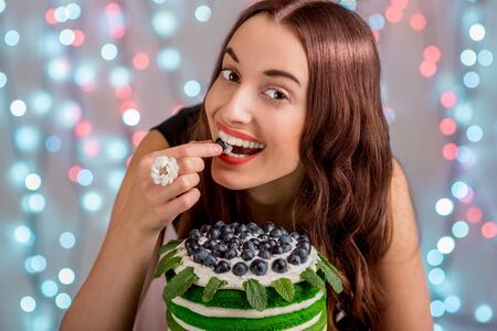 Beautiful girl with happy birthday cake tasting the grape on festive light backgroundの写真素材