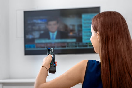 Young woman watching news on television with remote sitting on the sofa at home. Back view