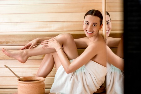 Young woman in white towel taking care of her legs sitting in Finnish saunaの写真素材