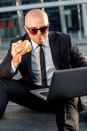 Businessman or banker working with laptop and having snack with burger outside the airport or contemporary buildingの写真素材