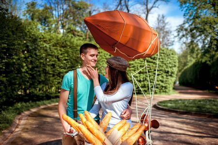 Lovely young couple riding and kissing on the bicycle with red dirigible and the basket full of baguettes in the park in the summerの写真素材