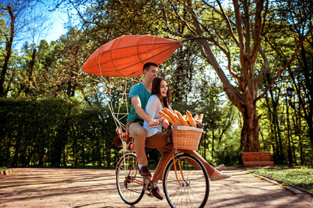 Lovely young couple riding bicycle with red dirigible and the basket full of baguettes in the park in the summerの写真素材