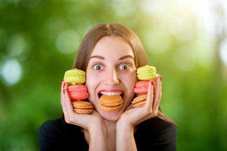 Young woman holding a bunch of colorful macaroons around her face and with on ein her mouse on blurred background, close-up planの写真素材