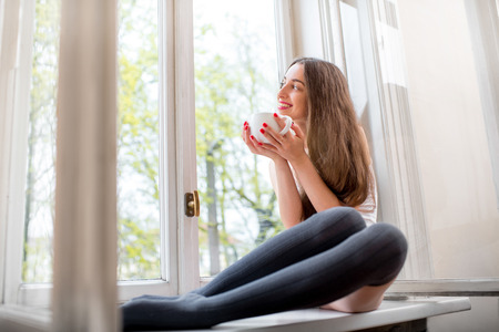Young and cute lady sitting on the windowsill and looking out the window with cup of coffee in the morningの写真素材