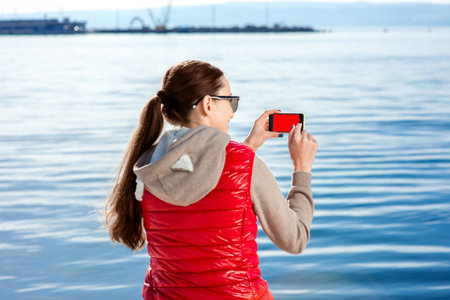 Sport woman dressed in warm clothes after the excercnise taking pictures with phone on the promenade...