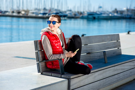 Sport woman dressed in warm clothes resting with phone on the bench near the sea in the spring