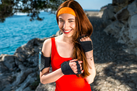 Sport woman in red shirt with sports gloves and phone holder looking at camera in the park near the seaの写真素材